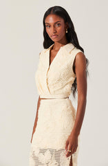 A woman is wearing a sleeveless, cream-colored textured suit that includes a collared top with a front opening and a high-waisted skirt. The fabric features intricate woven patterns, giving the outfit a sophisticated and elegant appearance.