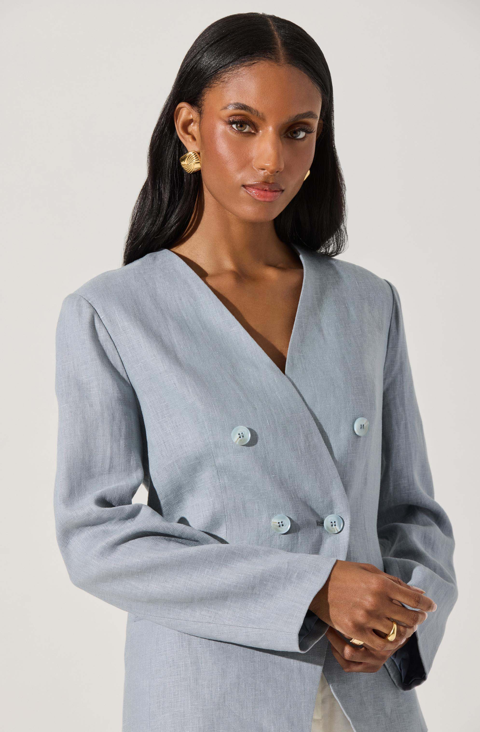 A woman is wearing a light gray blazer with a double-breasted design, featuring four buttons in two parallel rows. The blazer has a tailored fit with structured shoulders and long sleeves. She accessorizes with gold-tone, textured, hoop earrings and a gold ring on her right hand.