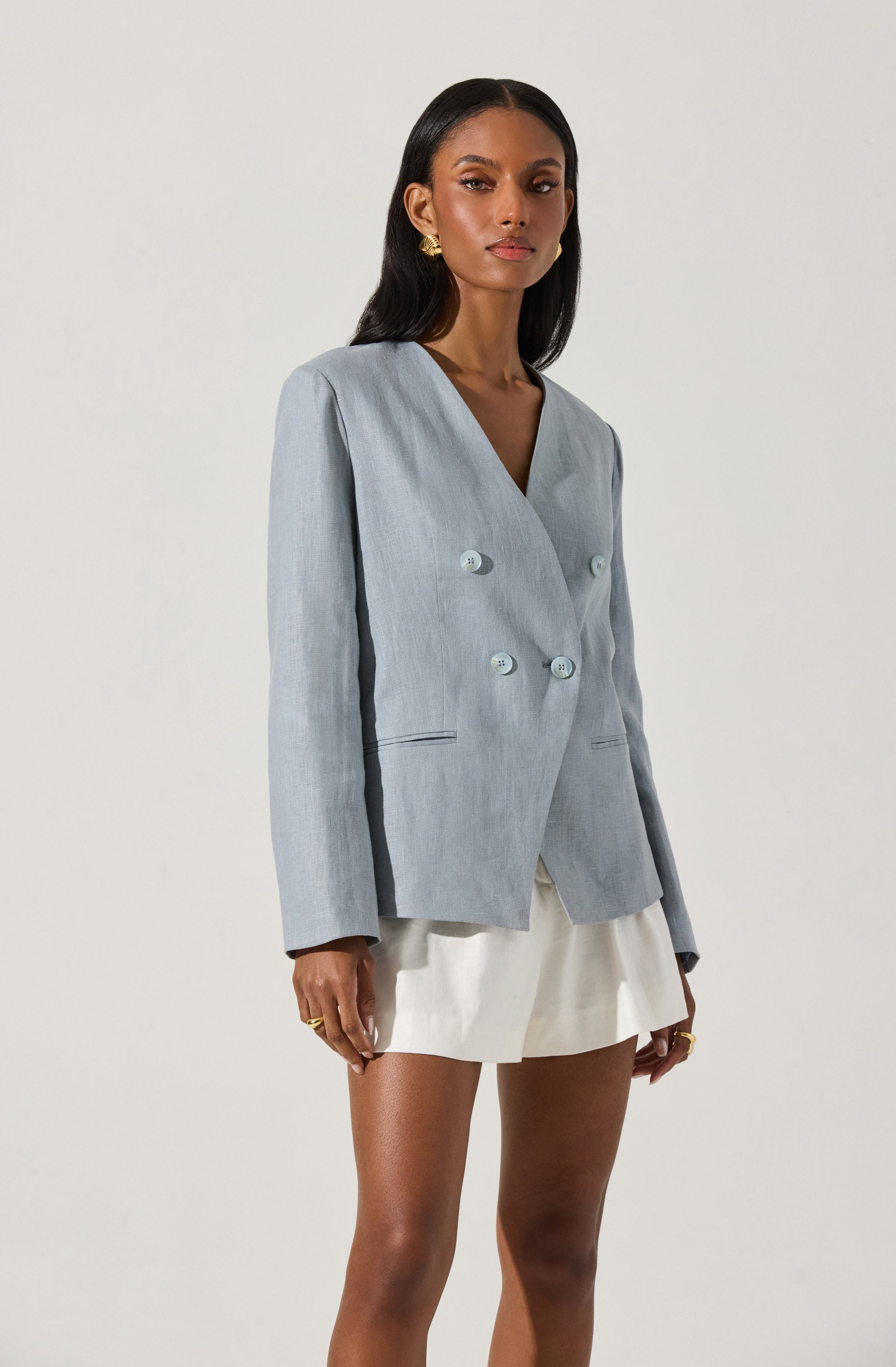 A woman is wearing a light gray, double-breasted blazer with four buttons, two on each side, and a notched collar. The blazer has a tailored fit with long sleeves and subtle slit pockets. She pairs it with a white mini skirt.