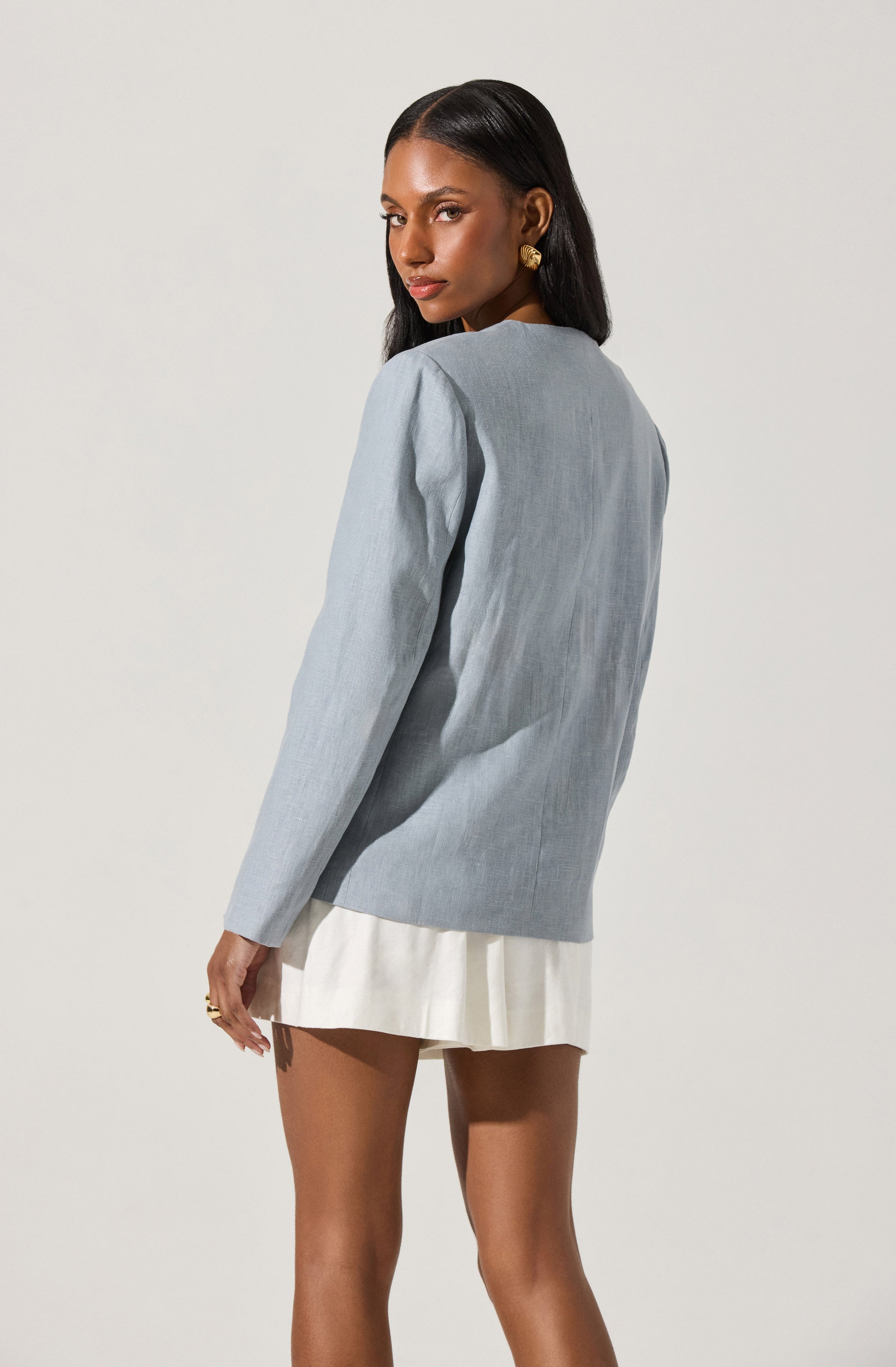 A woman with dark, straight hair is wearing a light gray, long-sleeved blazer-style top with minimal padding on the shoulders. She has paired it with a cream-colored, pleated mini skirt and is accessorized with gold earrings and a gold ring.
