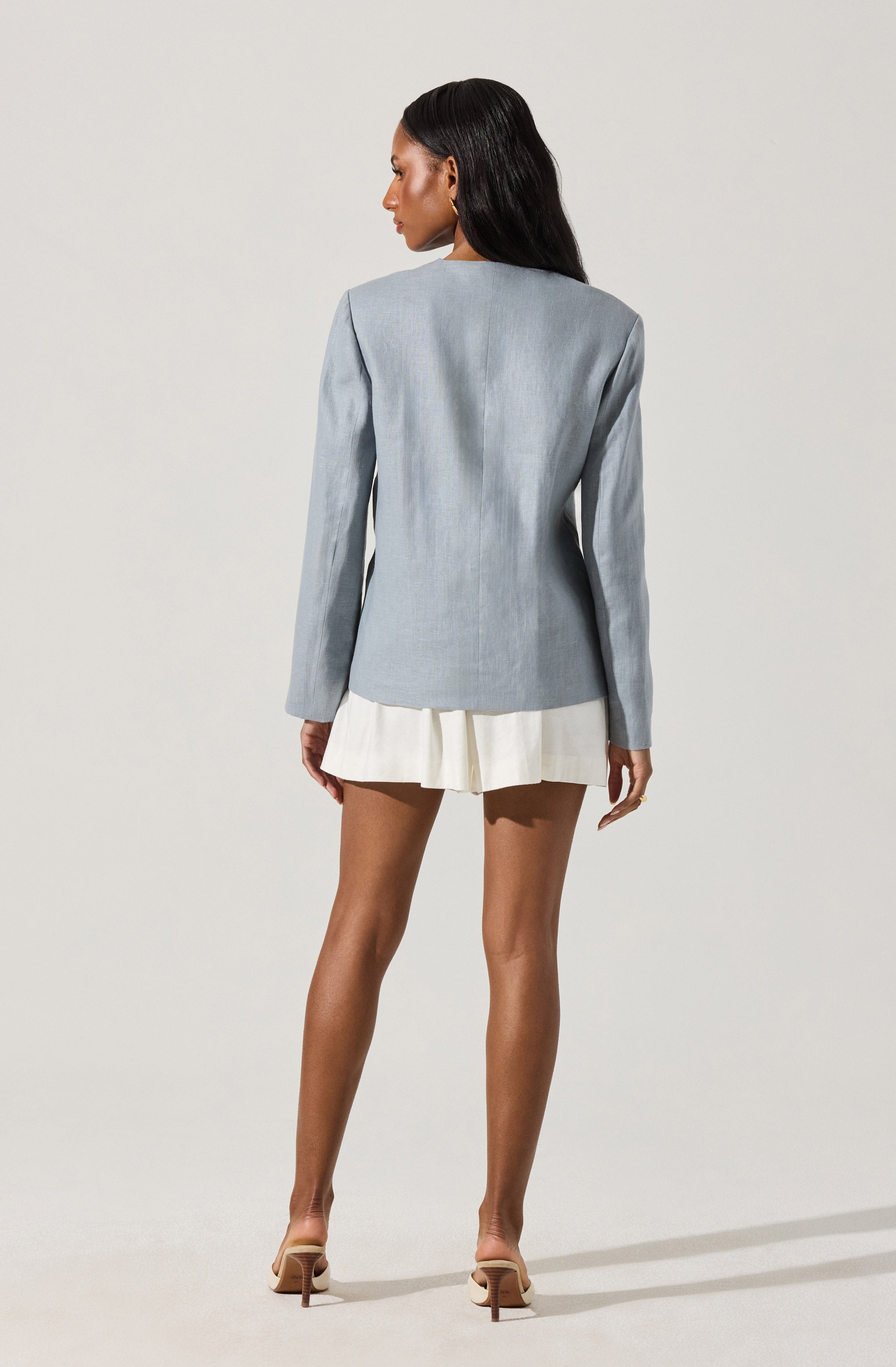 A woman is wearing a light gray blazer with a fitted silhouette and long sleeves. She pairs it with a short, white pleated skirt and beige high-heeled shoes. The outfit is stylish and sophisticated, suitable for a professional or semi-formal setting.