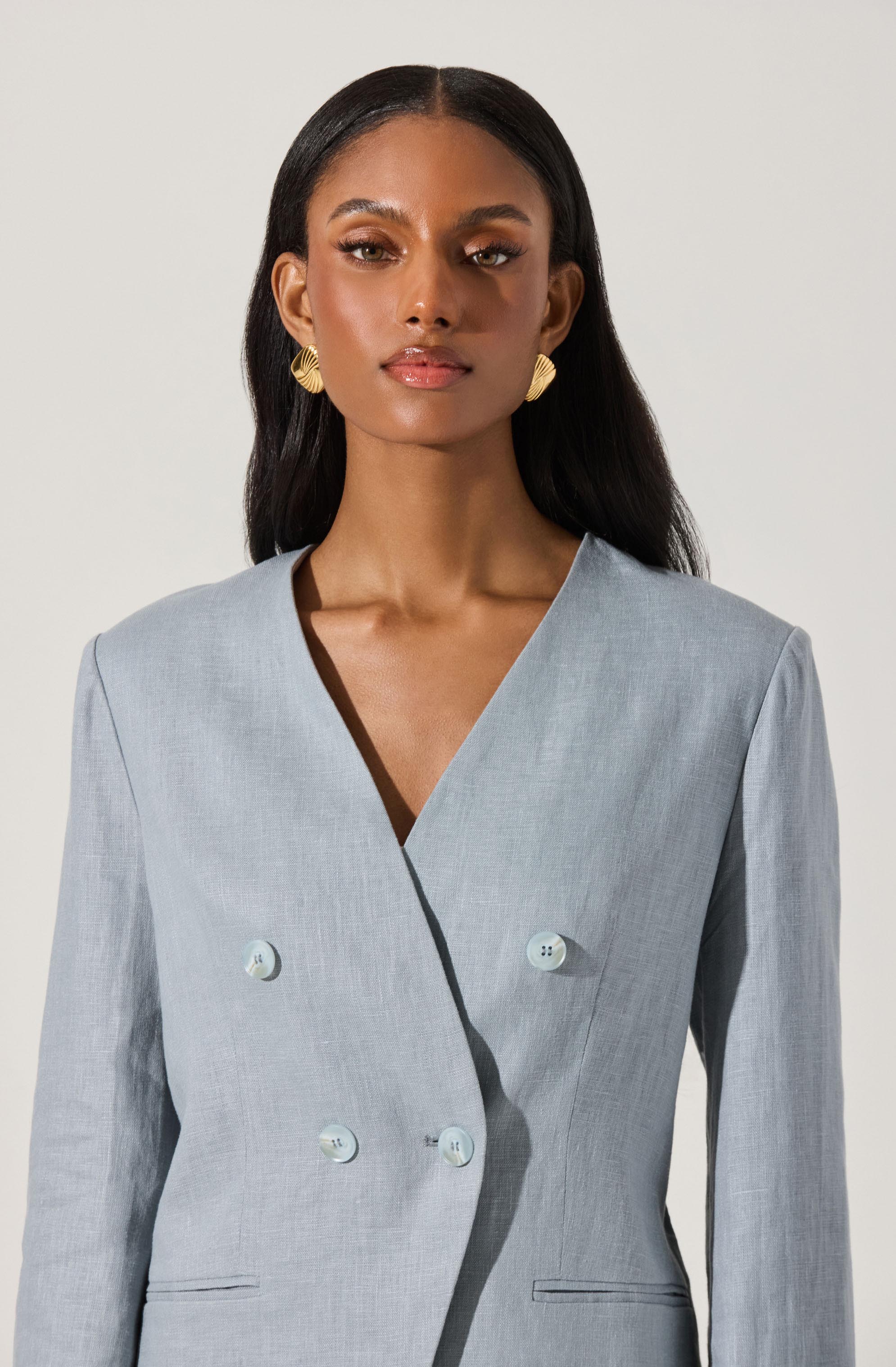 A woman is wearing a light gray, double-breasted blazer with six buttons on the front, three on each side. The blazer has a V-shaped neckline and appears to be made of a smooth, lightly textured fabric. She is accessorized with large gold, leaf-shaped earrings, and her hair is worn long, straight, and parted in the middle.