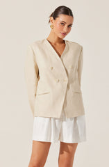 A woman is wearing a light-colored, double-breasted blazer with large buttons and two small, horizontal slit pockets. She is also dressed in matching light-colored high-waisted shorts. The outfit has a neutral, beige tone, and she accessorizes with gold earrings.