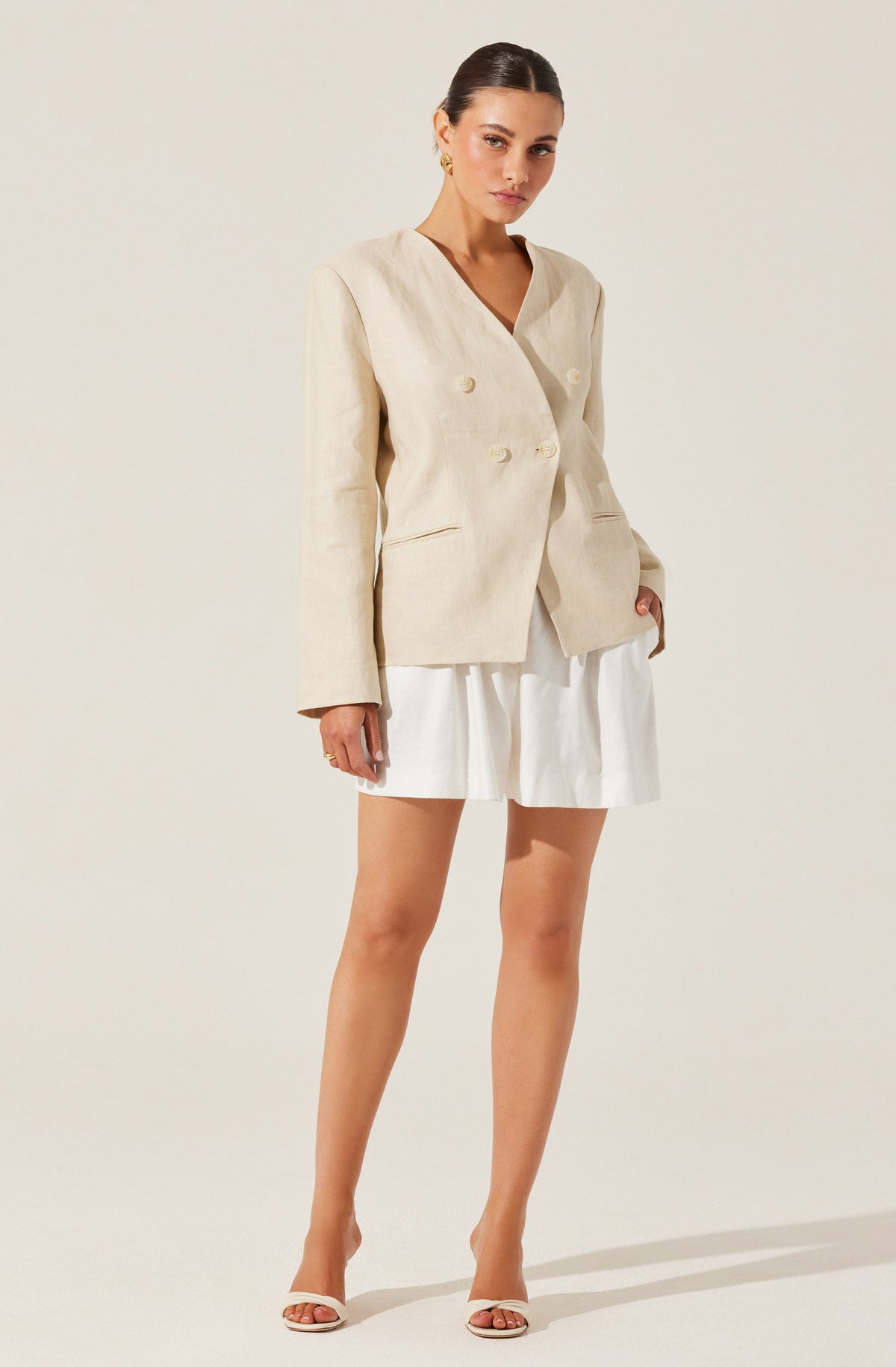 A woman is wearing a beige, double-breasted blazer with long sleeves and a V-shaped neckline. She pairs it with a white, pleated mini skirt and white high-heeled sandals.