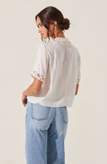 A woman is wearing a white short-sleeved blouse with lace trim on the sleeves and collar. She pairs it with light blue, high-waisted jeans. Her hair is styled in a low, loose bun.