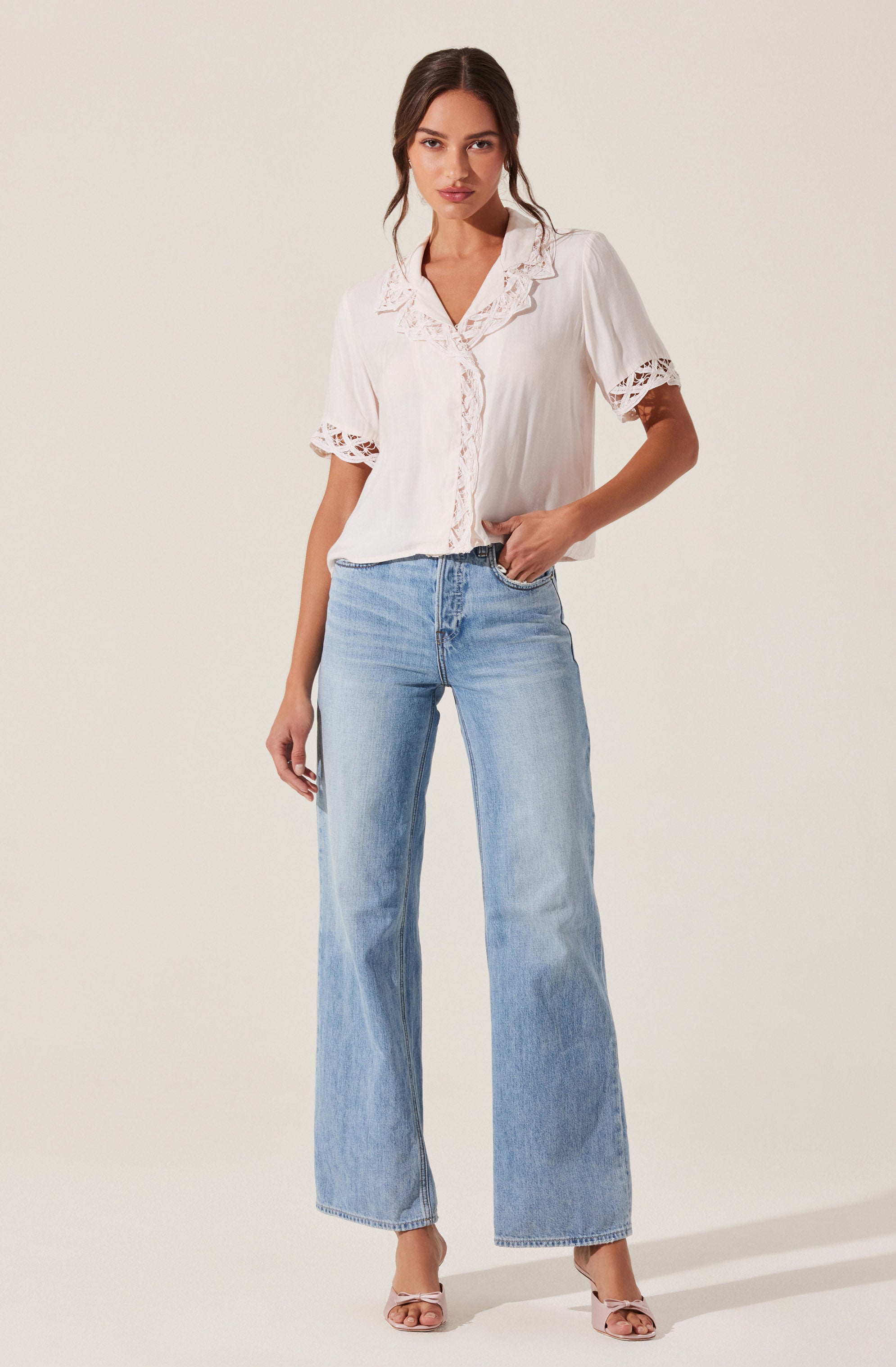 A woman is wearing a white short-sleeved blouse with lace details on the sleeves, collar, and front placket. The blouse has a slightly loose fit. She is also wearing light blue high-waisted, wide-leg jeans and beige strappy heels.