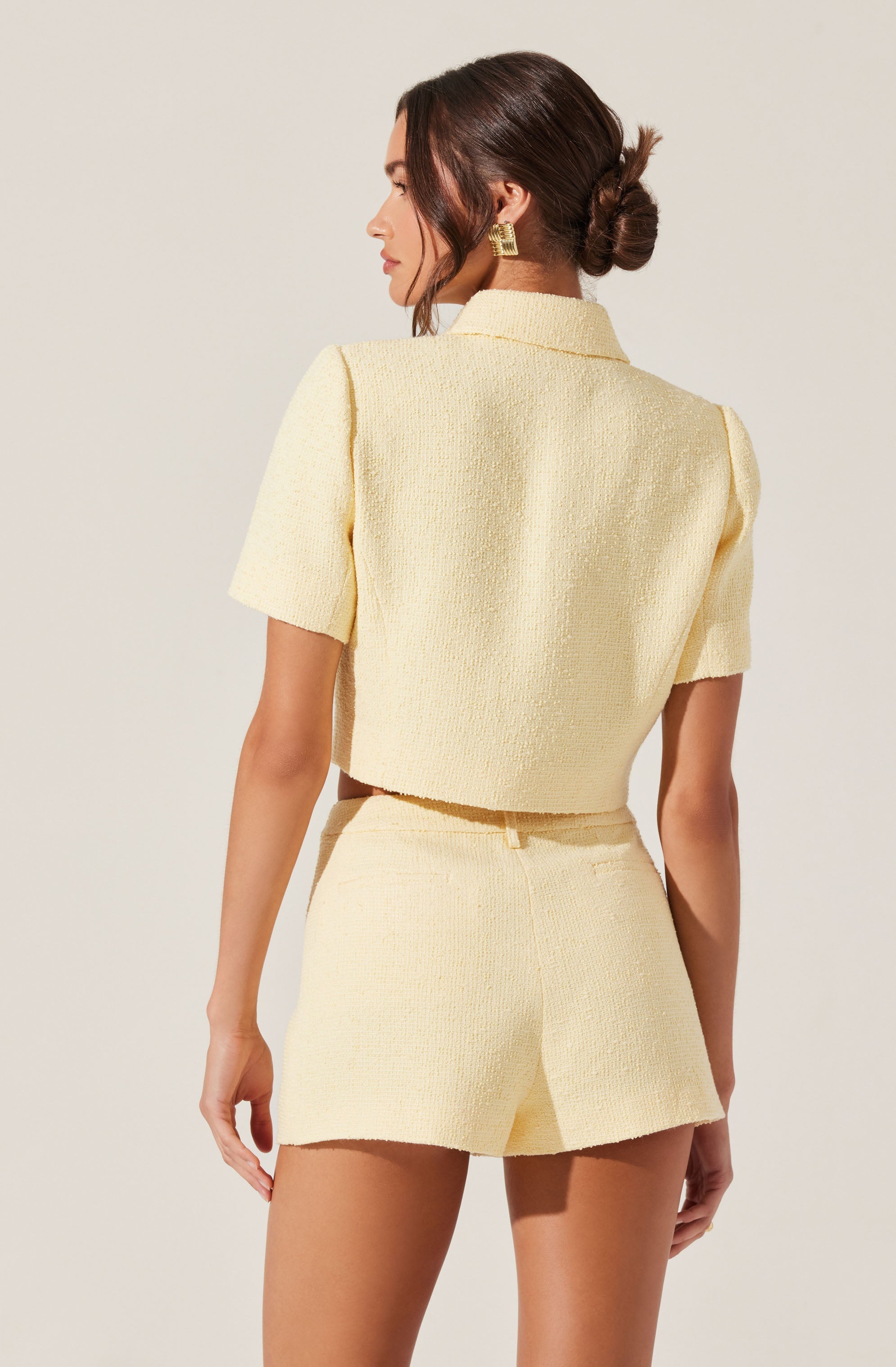 A woman is wearing a light yellow textured short-sleeve jacket with a collar, paired with matching high-waisted shorts. The outfit has a soft, woven appearance, and she accessorizes with large gold geometric earrings.
