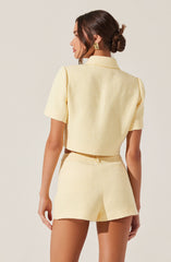 A woman is wearing a light yellow textured short-sleeve jacket with a collar, paired with matching high-waisted shorts. The outfit has a soft, woven appearance, and she accessorizes with large gold geometric earrings.