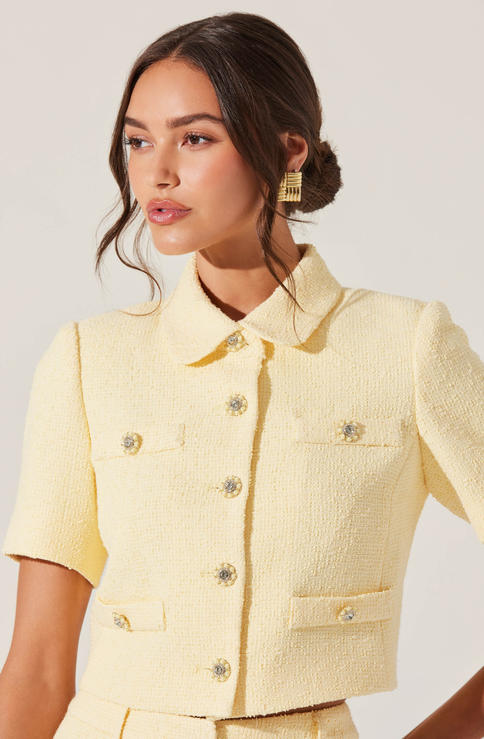A woman is wearing a light yellow, textured short-sleeved cropped jacket with a collar and metallic buttons. The jacket features a structured design with decorative pockets on the chest and waist.