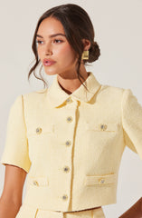 A woman is wearing a light yellow, textured short-sleeved cropped jacket with a collar and metallic buttons. The jacket features a structured design with decorative pockets on the chest and waist.