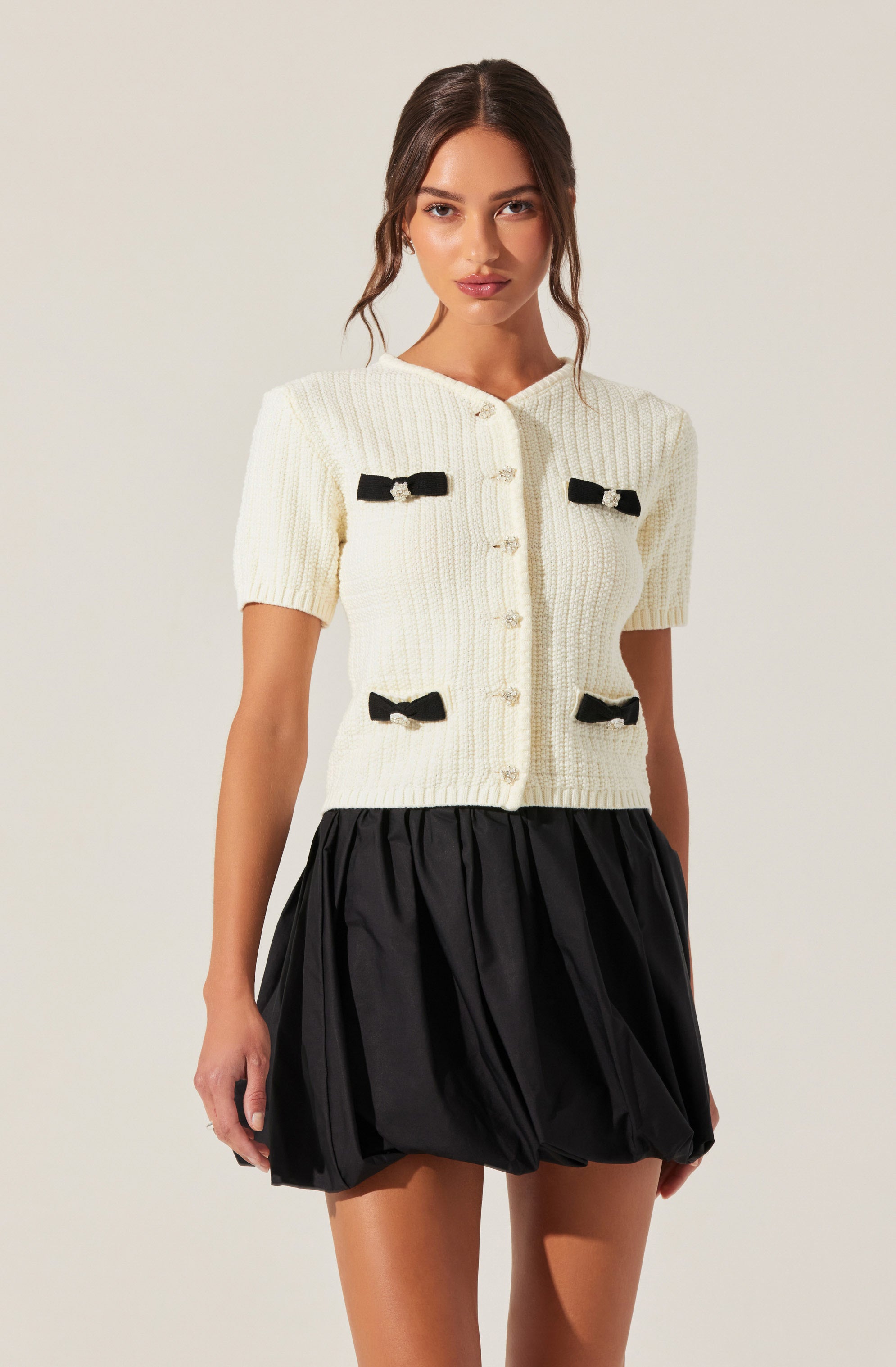 A woman is wearing a cream-colored, short-sleeved, knitted button-up jacket with small black bows on the front and near the pockets. She pairs it with a voluminous, black, bubble skirt that has a gathered, puffed appearance.