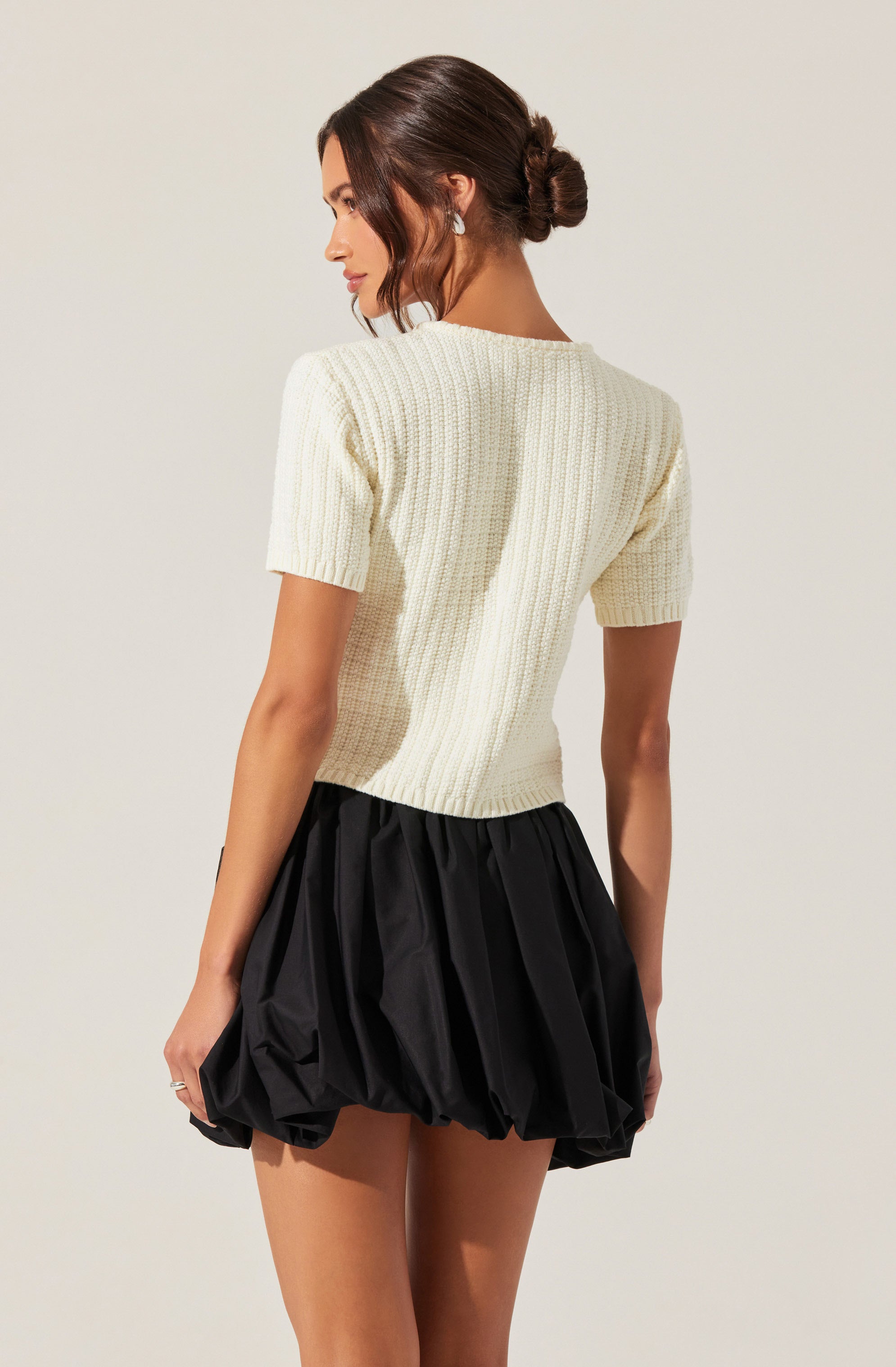 A woman is wearing a short-sleeved, cream-colored knit top with a textured, ribbed pattern. The top fits snugly and has a simple, classic design. She pairs it with a black, voluminous, balloon-style skirt that is gathered at the waist, creating a puffy appearance. The skirt's fabric appears slightly shiny and lightweight.