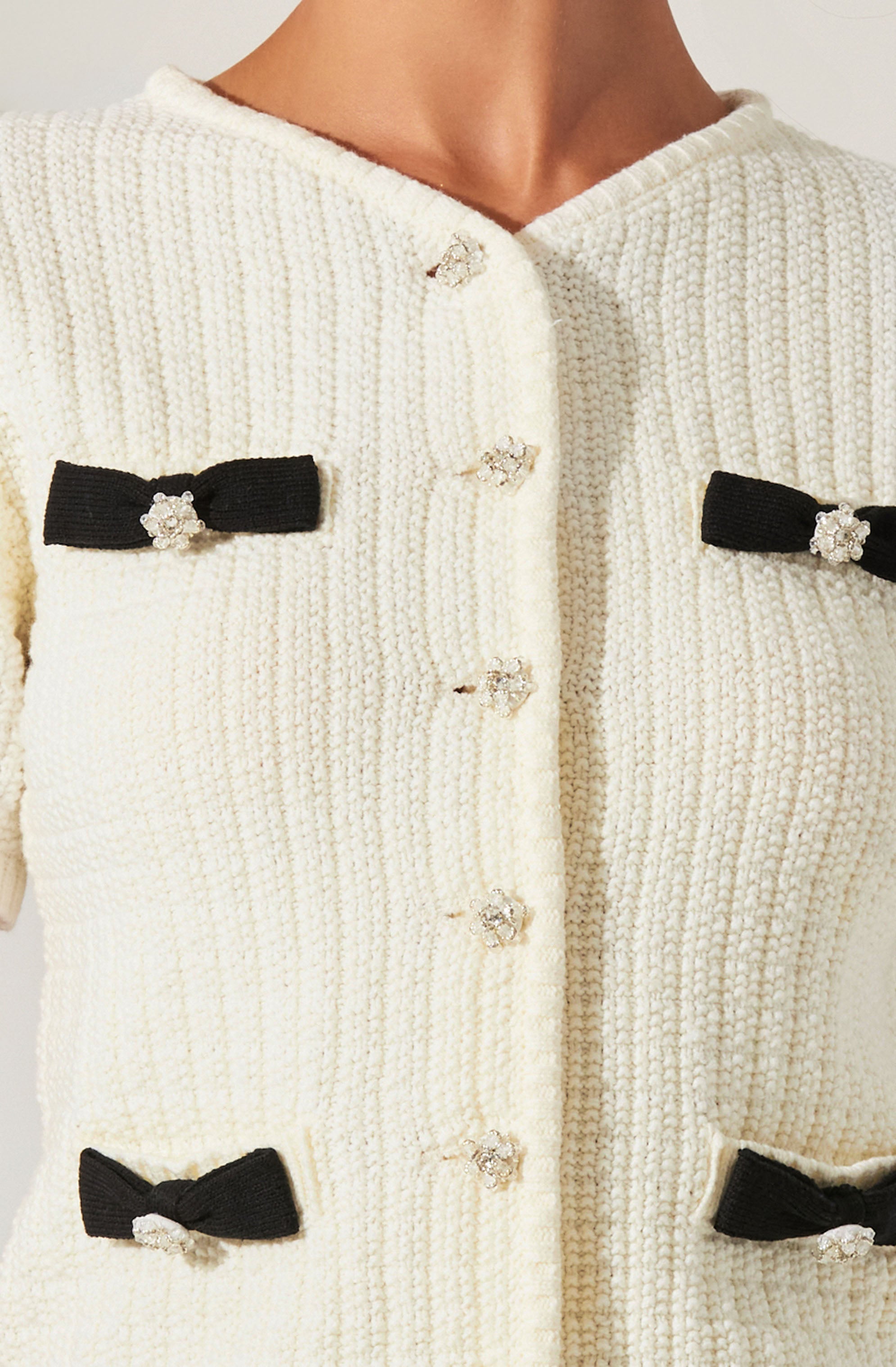 A cream-colored, textured knit cardigan with a rounded neckline. The cardigan features decorative black bows with small rhinestone accents positioned on the chest and near the waist, and it has a row of small, shiny buttons down the front.