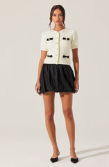A woman is wearing a short-sleeved, cream-colored, textured cardigan with black bow details on the front. She pairs it with a high-waisted, black, bubble skirt that is gathered and puffed at the bottom. She is also wearing black pointed-toe shoes with a bow detail on the front.