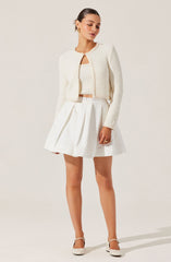A woman is wearing a white textured cropped cardigan with a round neckline and long sleeves. She pairs it with a matching white knit top underneath. The outfit is completed with a high-waisted white pleated skirt that falls above the knees, and white heels with ankle straps.