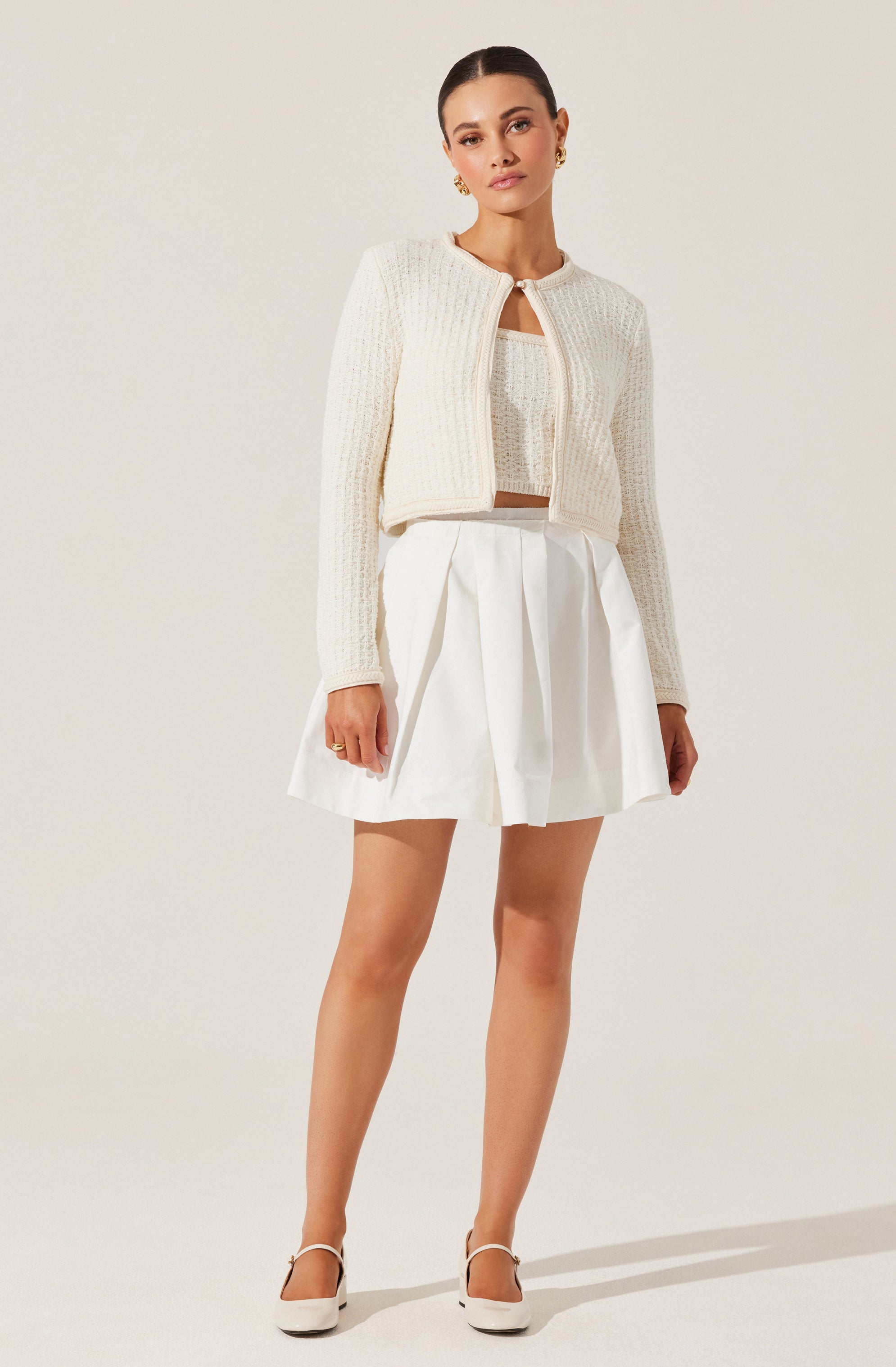 A woman is wearing a textured, cream-colored cardigan with a round neckline and an open front. Underneath, she has on a matching crop top. She pairs this with a high-waisted, white pleated skirt that falls above the knees. She completes the outfit with white high-heeled shoes with ankle straps.