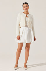 A woman is wearing a textured, cream-colored cardigan with a round neckline and an open front. Underneath, she has on a matching crop top. She pairs this with a high-waisted, white pleated skirt that falls above the knees. She completes the outfit with white high-heeled shoes with ankle straps.