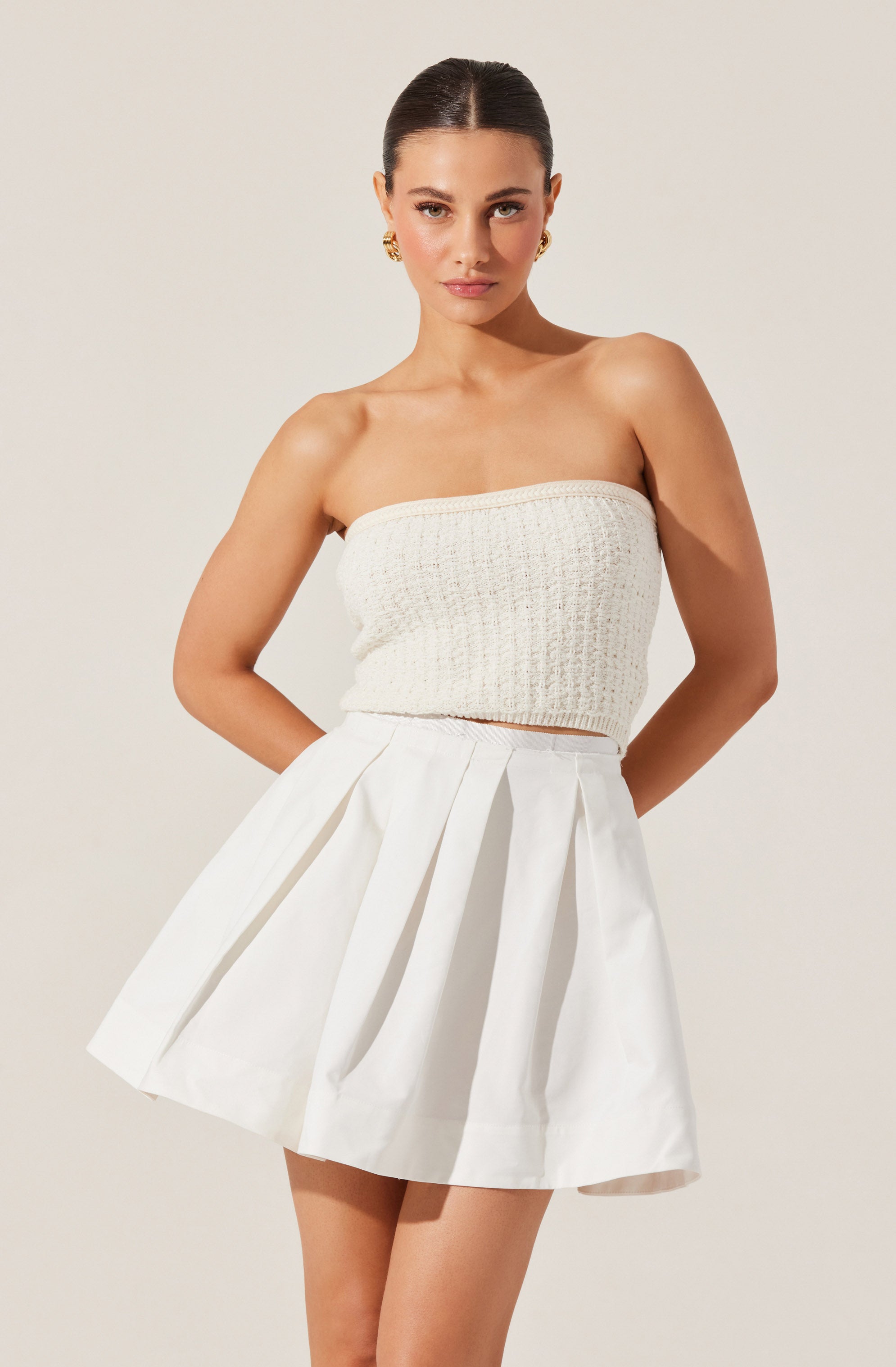 A woman is wearing a strapless, knitted, cream-colored top paired with a high-waisted, white pleated skirt. She has her hair styled in a sleek, pulled-back look and is accessorized with gold earrings.