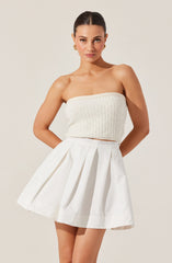 A woman is wearing a strapless, knitted, cream-colored top paired with a high-waisted, white pleated skirt. She has her hair styled in a sleek, pulled-back look and is accessorized with gold earrings.