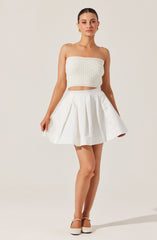 A woman is wearing a strapless, cream-colored, textured crop top with a banded elastic top edge. She pairs it with a high-waisted, white, pleated skirt that has a wide waistband and features box pleats for added volume. She completes the outfit with cream-colored closed-toe heels.