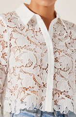 A white, long-sleeved button-up shirt featuring an intricate, floral lace pattern throughout. The shirt has a classic pointed collar and white buttons down the front. The lace design varies with leaves and flowers, creating a semi-transparent look. The shirt is paired with light blue jeans.