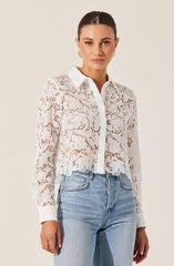 A woman is wearing a white lace long-sleeve button-up shirt with a floral pattern and a classic collar. She pairs it with light-wash blue jeans. She has dark hair pulled back, gold earrings, and a neutral expression.