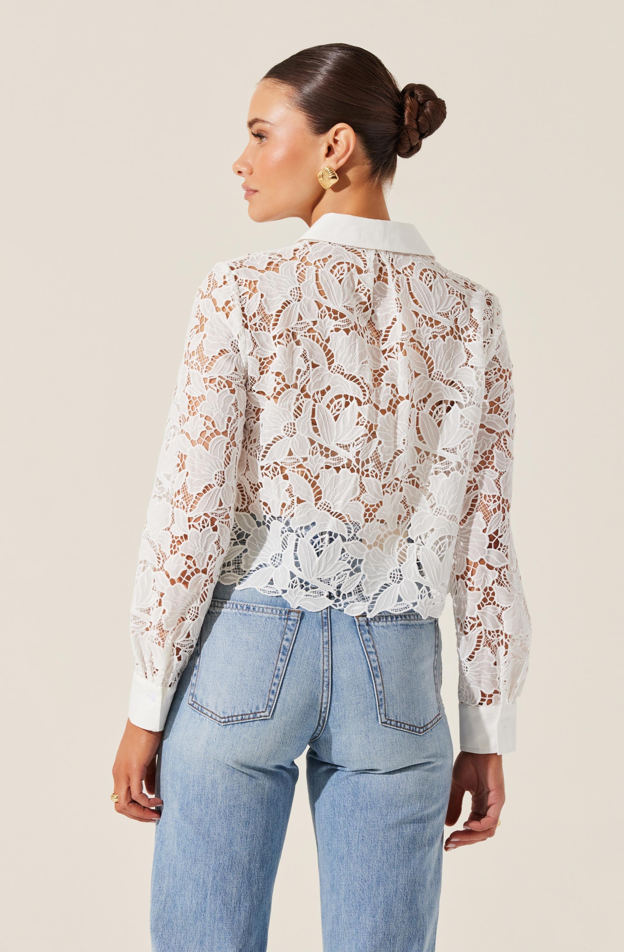 A woman is wearing a long-sleeve, white lace top with a floral pattern. The top has a solid white collar and cuffs. She is also wearing light-wash denim jeans with prominent back pockets.