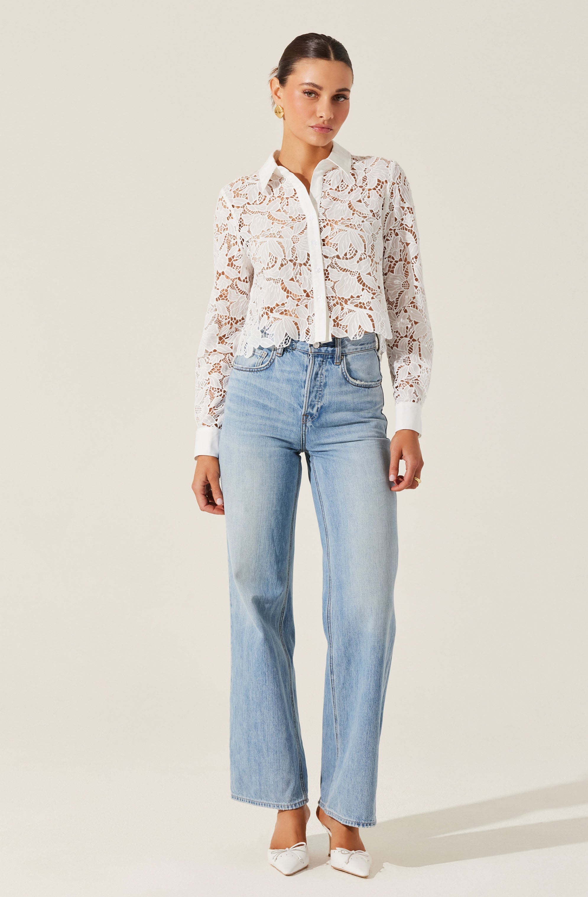 A woman is wearing a white lace long-sleeve shirt with a collar and a button-down front. The shirt features a floral lace pattern and is slightly cropped with a gathered hem. She is also wearing light blue high-waisted, straight-leg jeans with a relaxed fit, and white shoes with pointed toes.
