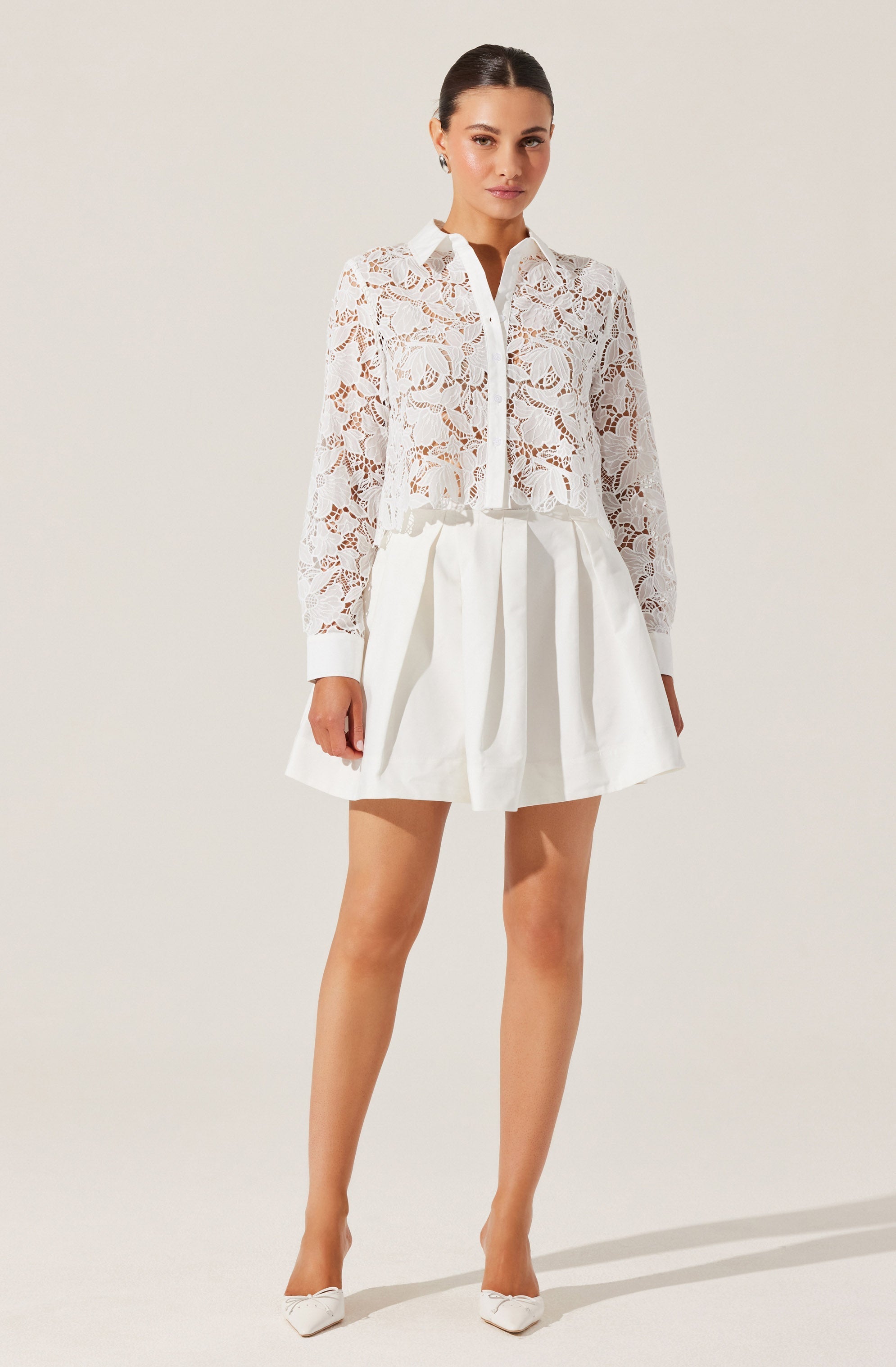A woman is wearing a white, long-sleeved, button-up blouse made of lace with a floral pattern. The blouse has a collar and is cropped, ending just above the waist. She is also wearing a white, high-waisted skirt with pleats or gathers, creating a voluminous, A-line silhouette. She completes the look with white pointed-toe high heels.