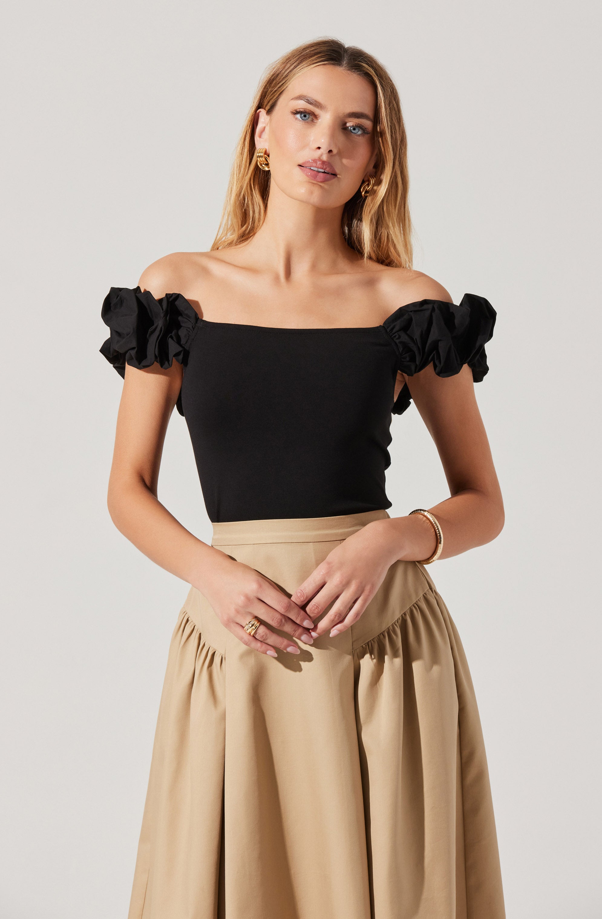 A woman is wearing an off-the-shoulder black top with puffy, gathered sleeves. She pairs it with a high-waisted, beige skirt that has a gathered, voluminous design.