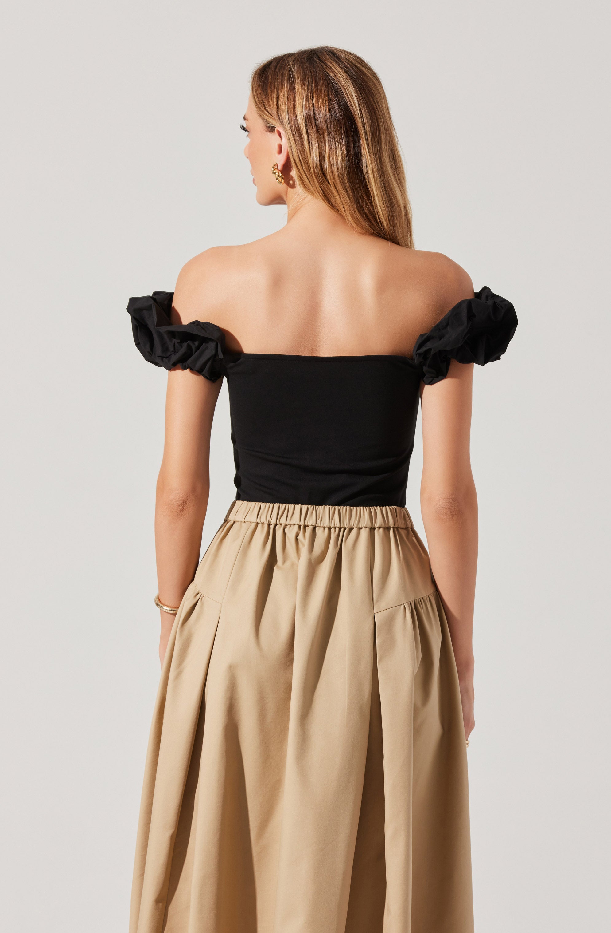 A woman is wearing a black off-the-shoulder top with puffed short sleeves and a pair of high-waisted, beige, wide-leg pants with an elastic waistband and gathered fabric. She accessorizes with gold hoop earrings and a gold bracelet.
