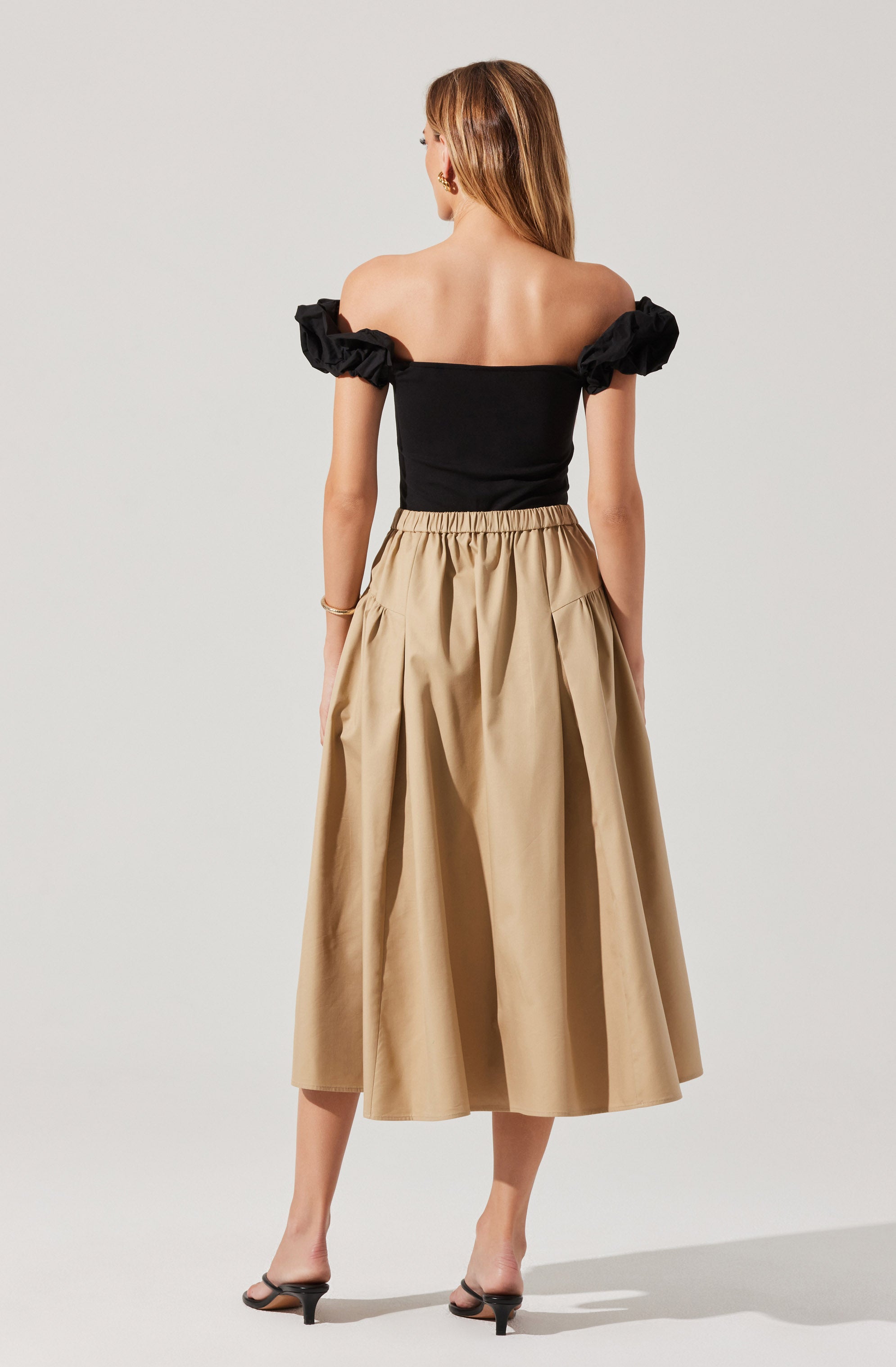 A woman is wearing a black off-the-shoulder top with ruffled short sleeves, paired with a high-waisted, beige, ankle-length skirt that has a gathered, pleated design. She is also wearing black open-toe heels.