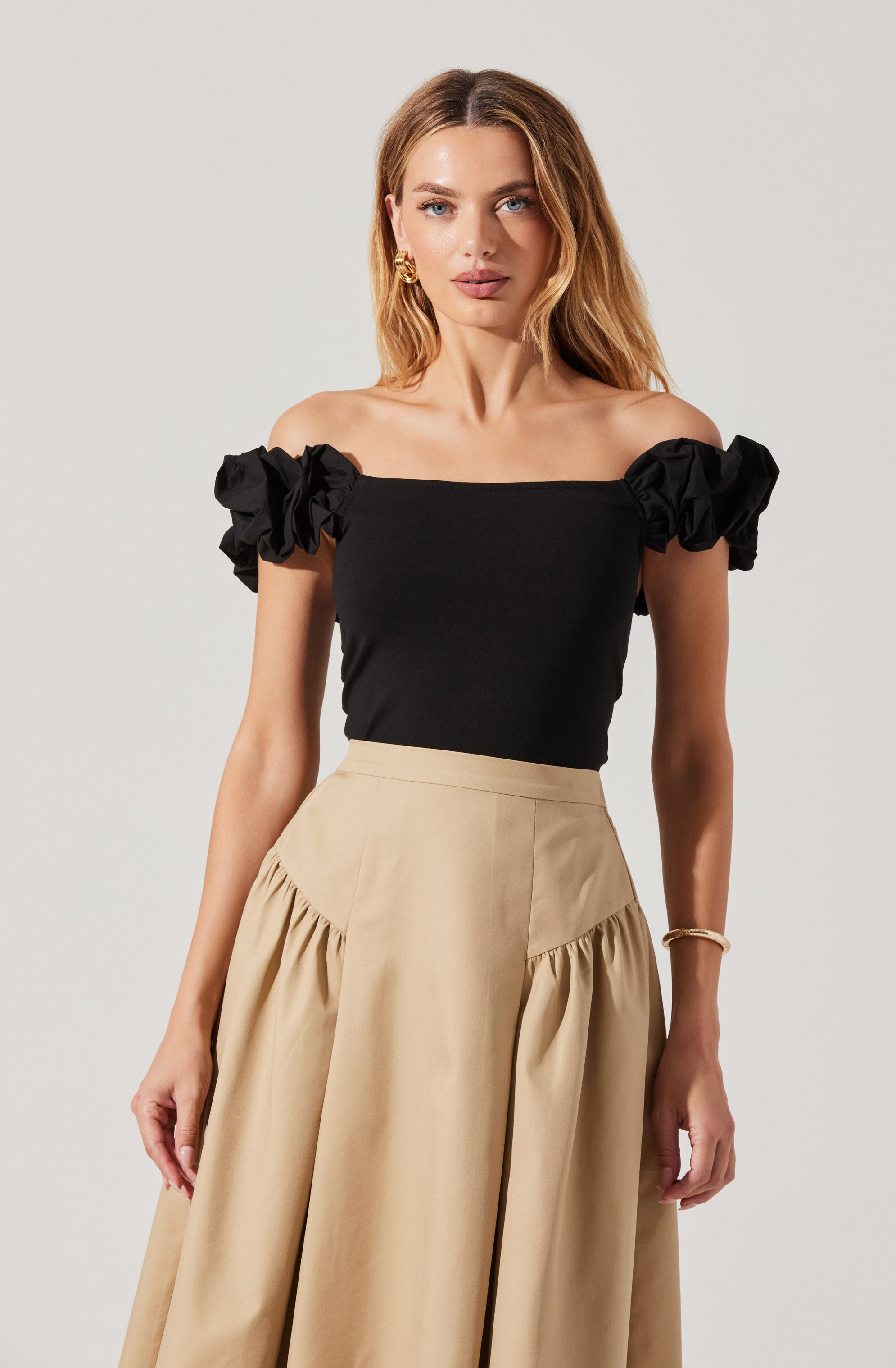 A woman is wearing a black off-the-shoulder top with ruffled short sleeves and a high-waisted beige skirt with gathered fabric details.