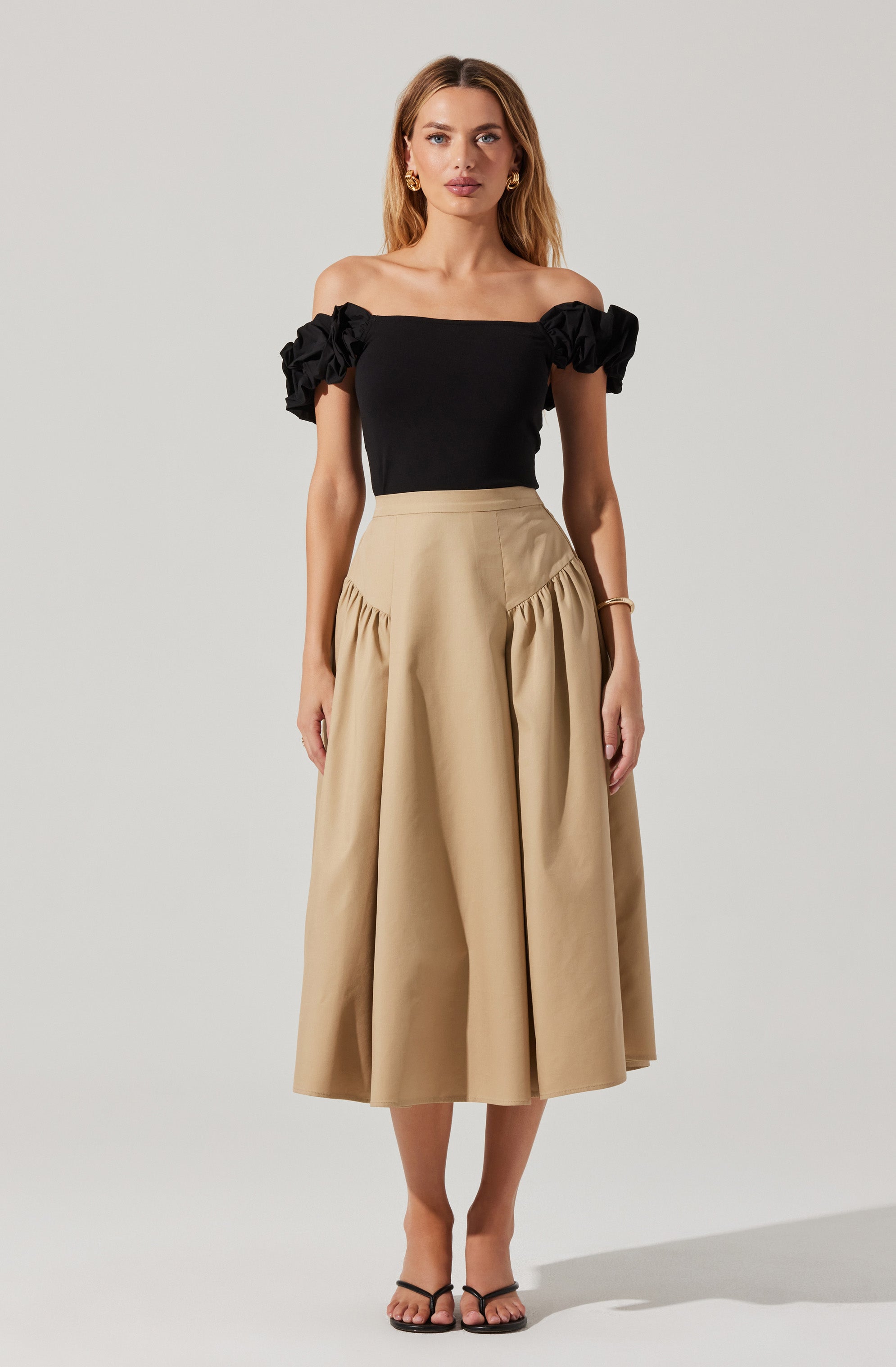 A woman is wearing a black off-the-shoulder top with ruffled sleeves and a beige, high-waisted, A-line midi skirt with pleats and gathered details on the sides. She is also wearing black strappy heels and gold jewelry.