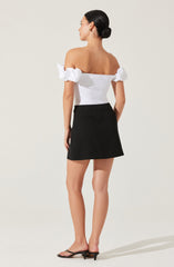 A woman is wearing an off-the-shoulder white top with puffed short sleeves and a fitted bodice, paired with a black mini skirt that has a subtle slit at the back and a small pocket on the right side. She is also wearing black open-toe heels with a small heel.