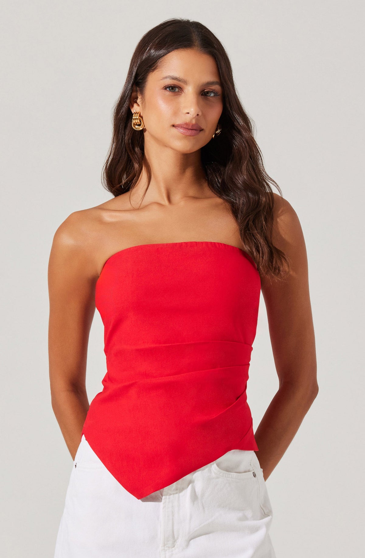 A woman with long, wavy brown hair is wearing a strapless red top that fits snugly around her torso. She has on large, gold hoop earrings and is dressed in white pants.