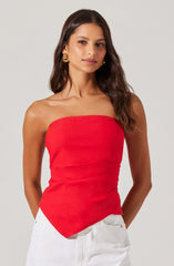 A woman with long, wavy brown hair is wearing a strapless red top that fits snugly around her torso. She has on large, gold hoop earrings and is dressed in white pants.