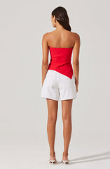 A woman is wearing a strapless, bright red top that fits snugly around her torso. She has paired it with high-waisted white shorts that have large front pockets and a loose fit. She is also wearing nude-colored, open-toe heels.