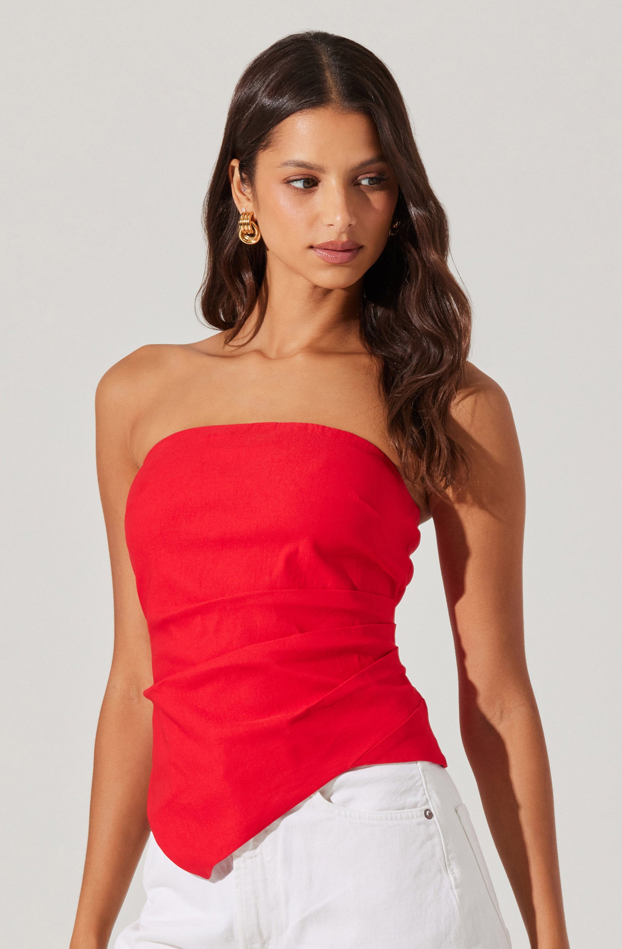 A woman is wearing a strapless red top that fits snugly and has a slightly asymmetrical hemline. She pairs it with white pants.