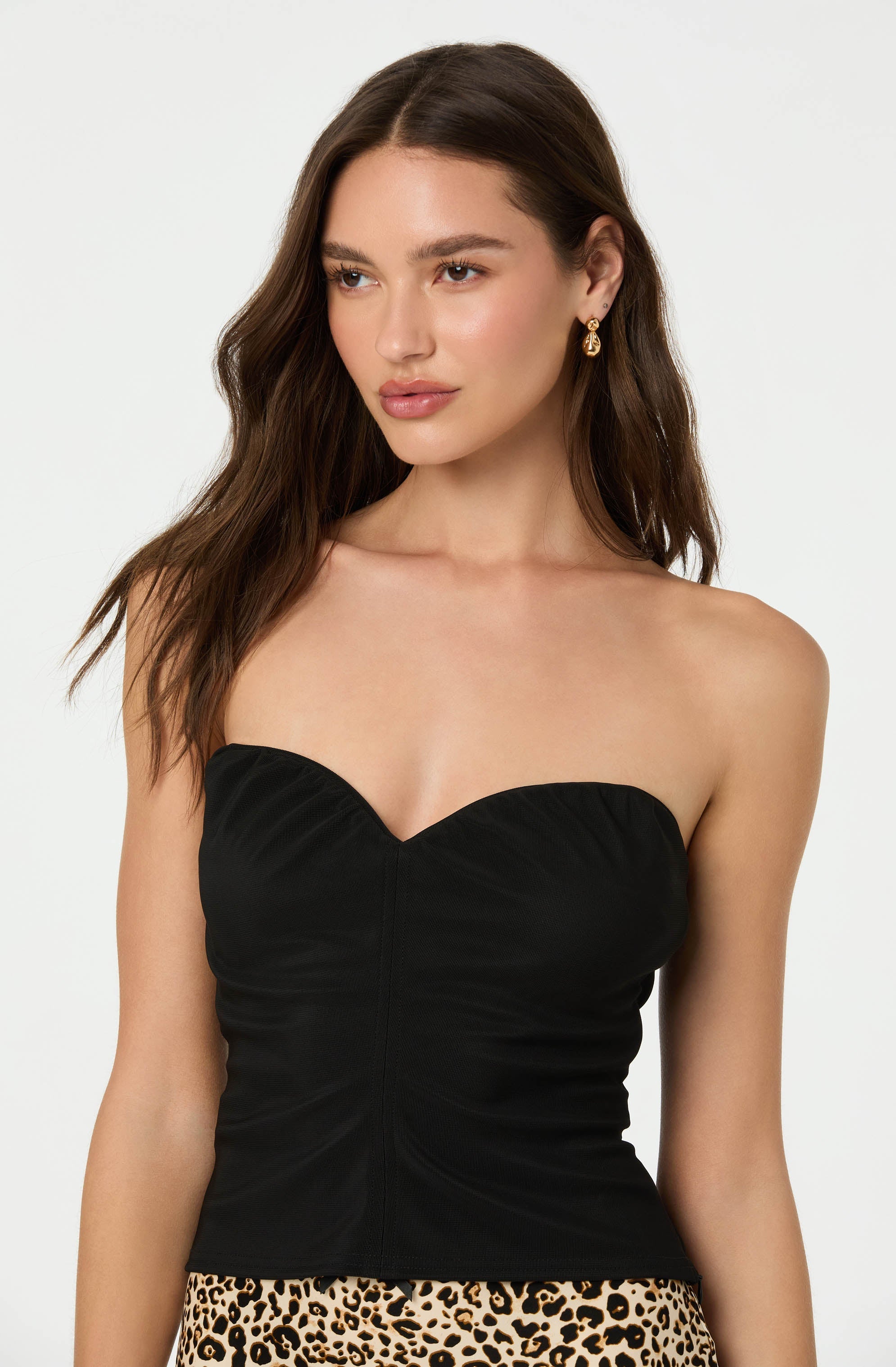 A young woman is wearing a strapless black top with a sweetheart neckline. She is also wearing gold drop earrings and a leopard print skirt.