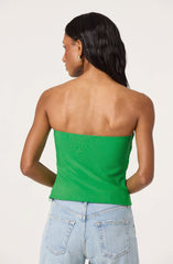 The image shows a woman wearing a bright green strapless top that fits snugly around her torso. She is also wearing light blue, high-waisted jeans with visible back pockets. The woman has long, dark, wavy hair and is accessorized with gold hoop earrings.