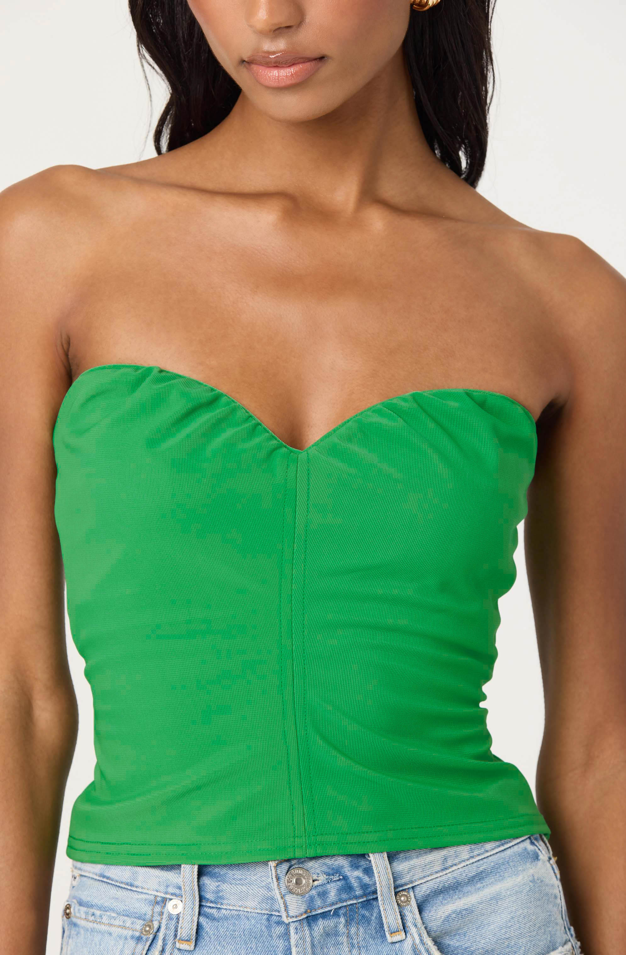 A woman is wearing a strapless, fitted green bustier top with a center seam detail. The top is smooth and slightly ruched on the sides. She is also wearing light-wash denim jeans.
