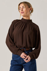 A woman is wearing a long-sleeve, dark brown blouse with a high, gathered neckline. The blouse has puffy sleeves that are gathered at the cuffs. She is also wearing blue jeans with a brown belt and hoop earrings.