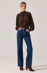A woman is wearing a dark brown long-sleeve blouse with a high, gathered neckline and billowy sleeves that are cinched at the wrists. She pairs the blouse with high-waisted, straight-leg blue jeans secured with a brown belt, and completes the outfit with brown heeled ankle boots.