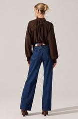 A woman is wearing a loose-fitting, long-sleeved chocolate brown blouse with gathered fabric at the neckline and cuffs. She pairs it with high-waisted, wide-leg blue jeans and brown heeled shoes. Her hair is styled in a neat bun.