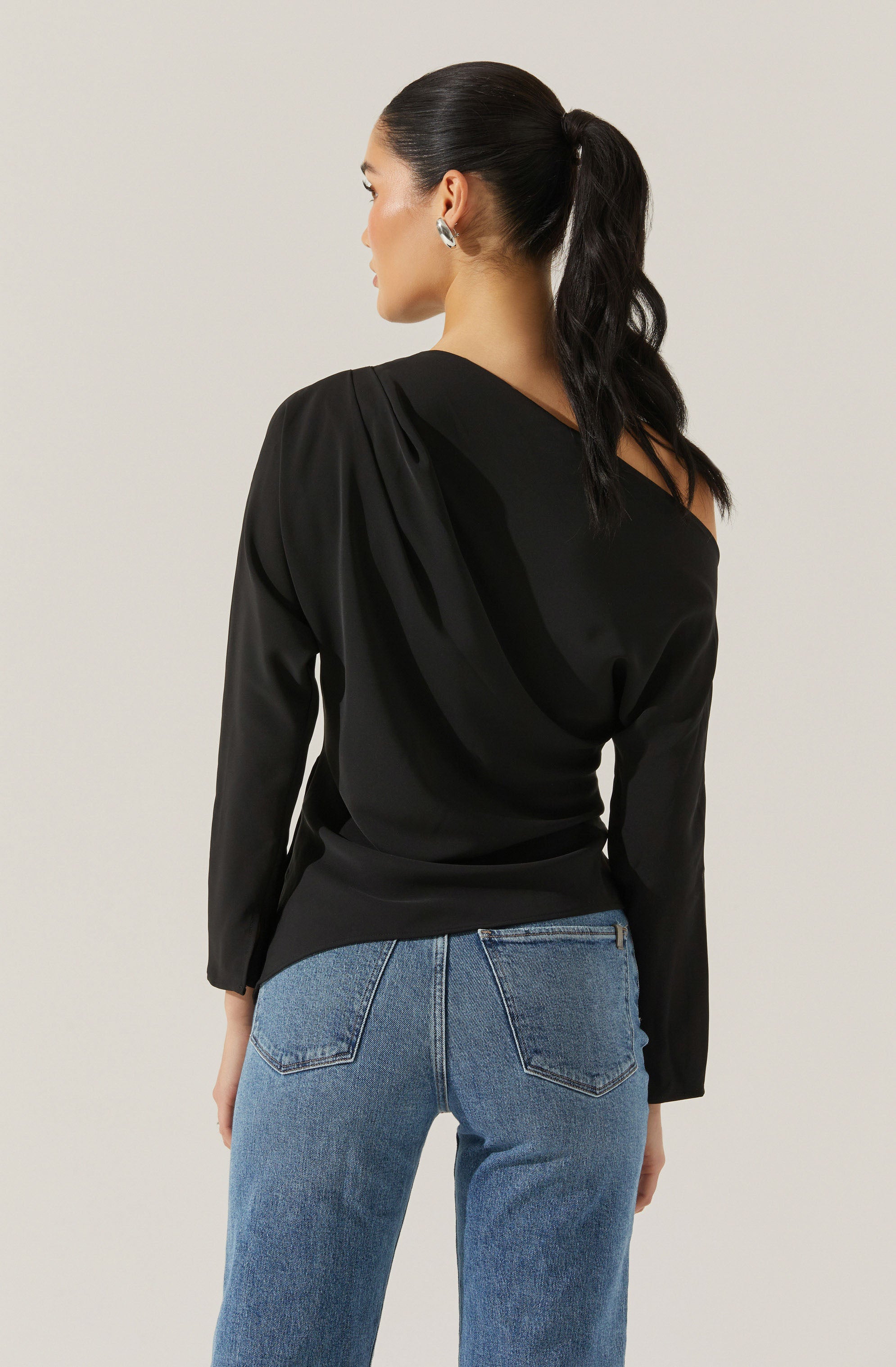 A woman is wearing a black long-sleeve top with a one-shoulder design and pleated detailing on the shoulder. She is also dressed in light blue high-waisted jeans with back pockets.