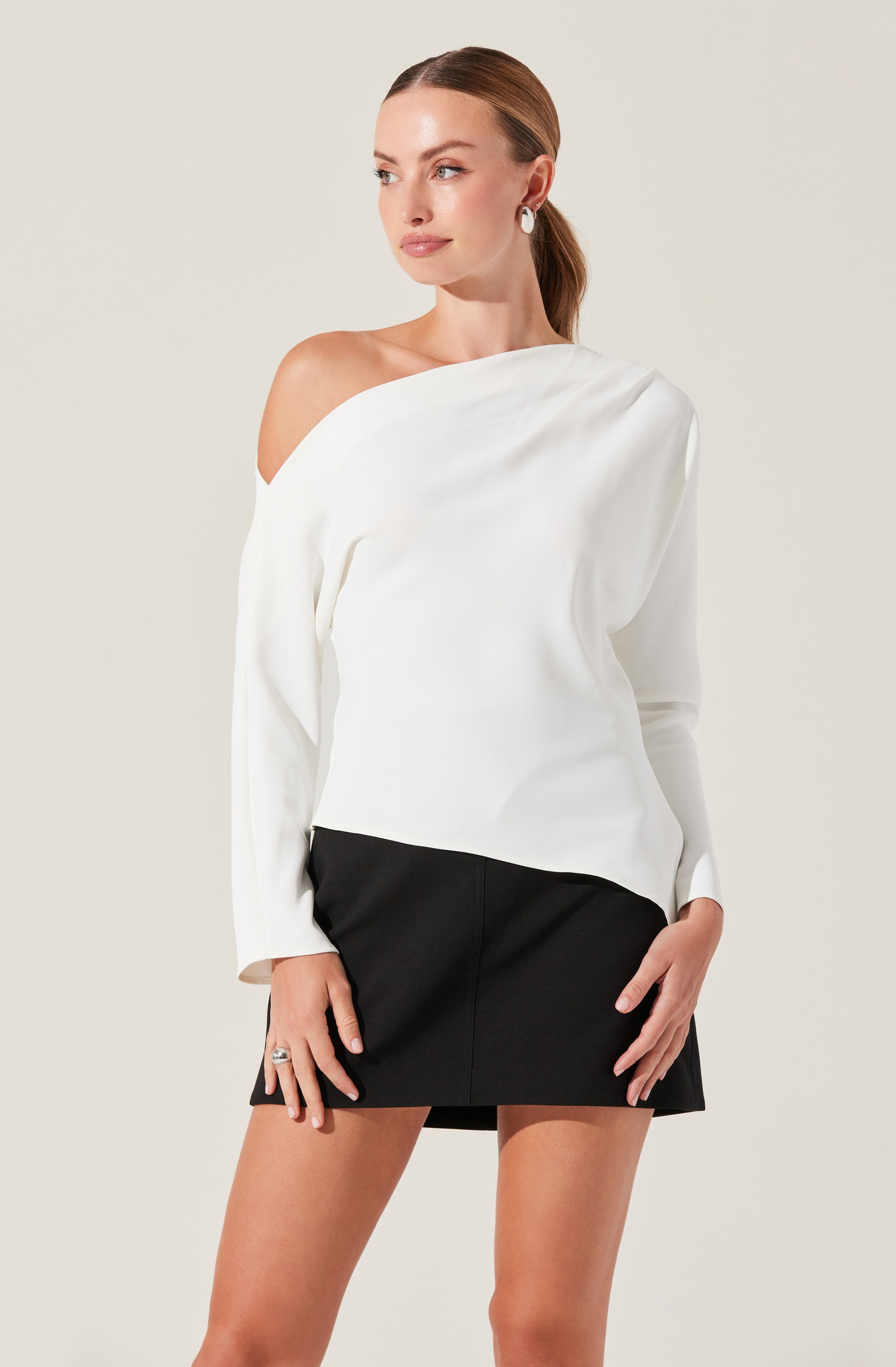 A woman is wearing a white, off-the-shoulder top with long sleeves, paired with a black mini skirt. The top has a relaxed fit with draped fabric, and the skirt is fitted and above the knee.