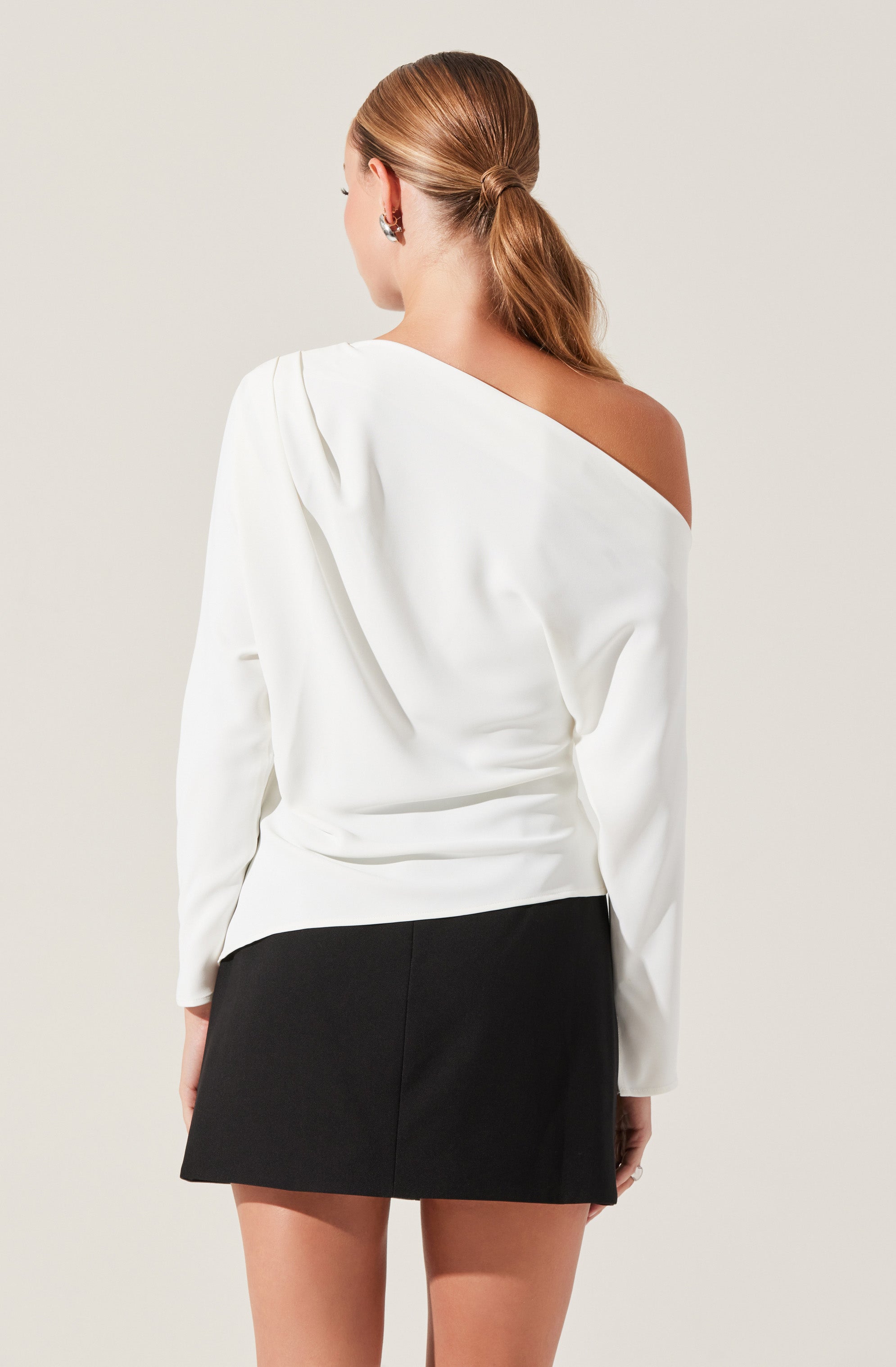 A woman is wearing an off-the-shoulder white top with long sleeves, paired with a black mini skirt. The top drapes elegantly, exposing one shoulder, while the skirt is fitted and short. Her hair is styled in a low ponytail.