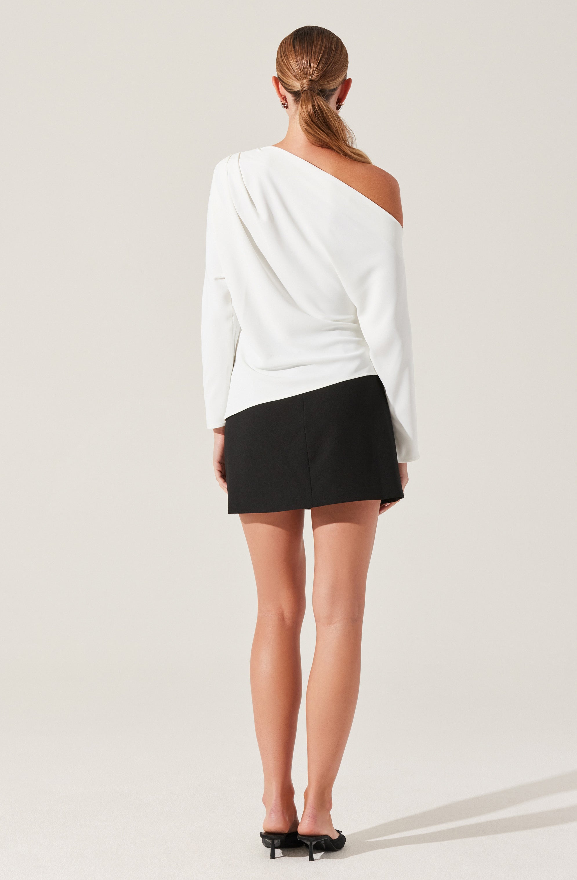 A woman is wearing a white, asymmetrical, off-the-shoulder top with one long sleeve and the other shoulder exposed. She has paired it with a black, high-waisted mini skirt and black heeled sandals. Her hair is styled in a low ponytail.