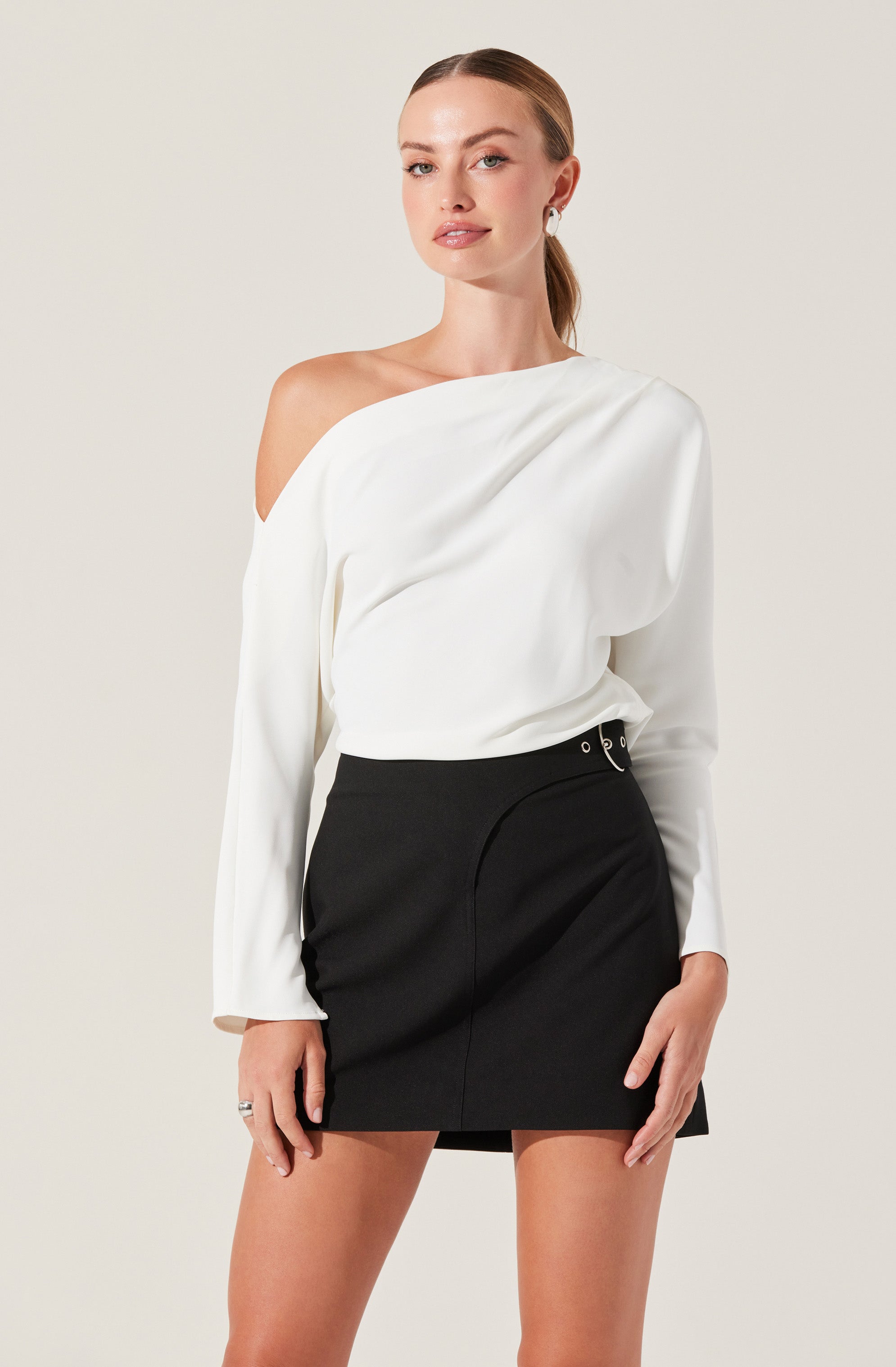 A woman is wearing a stylish, one-shoulder white blouse with voluminous long sleeves, paired with a high-waisted black skirt that features decorative grommets and a subtle front seam. She has her hair in a sleek ponytail and is accessorized with earrings and a ring.